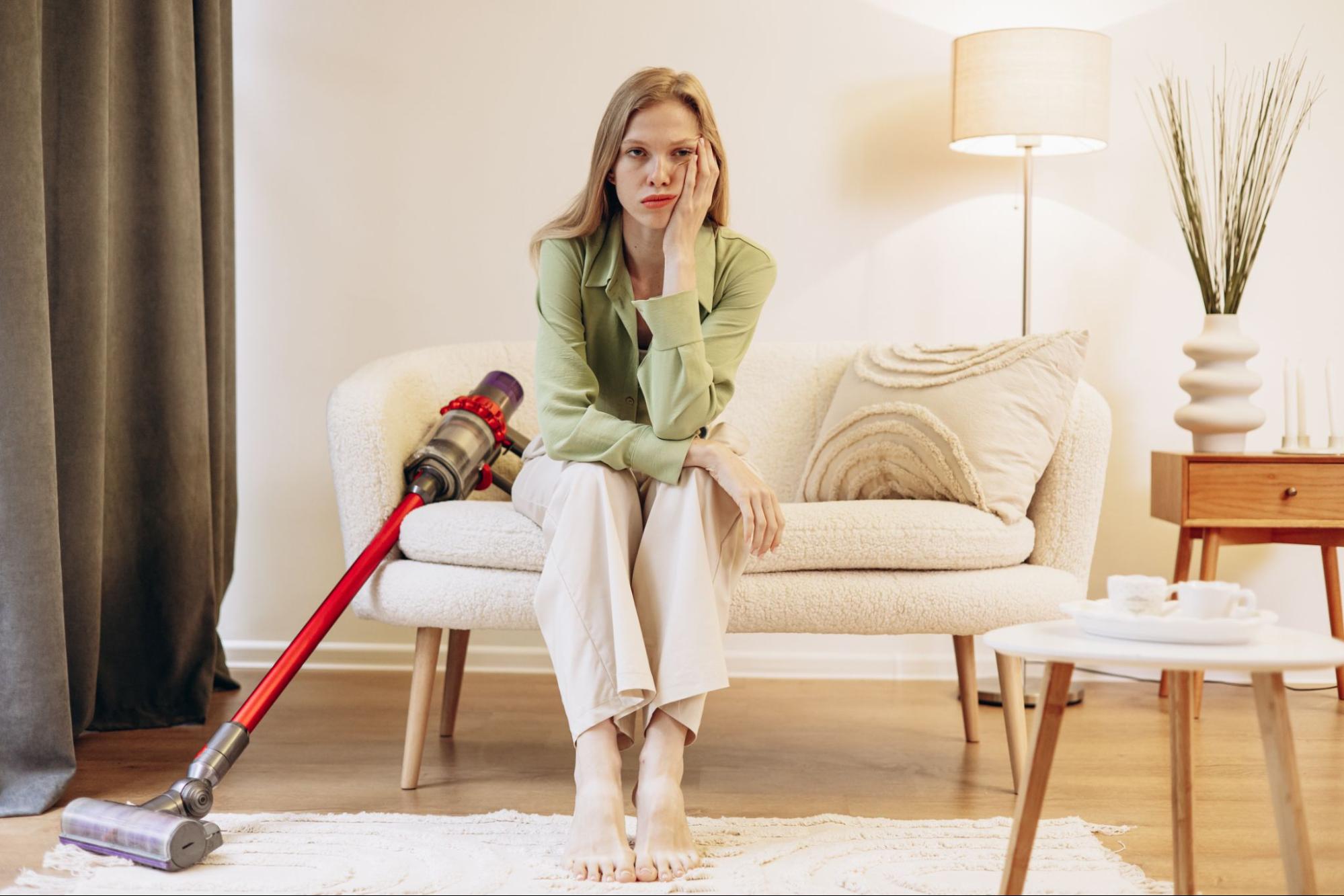 Uma mulher sentada em um sofá com um aspirador de pó do lado.